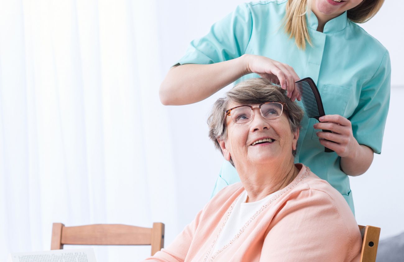 Eine Frau bürstet das Haar einer älteren Dame, während sie sich freundlich unterhalten.